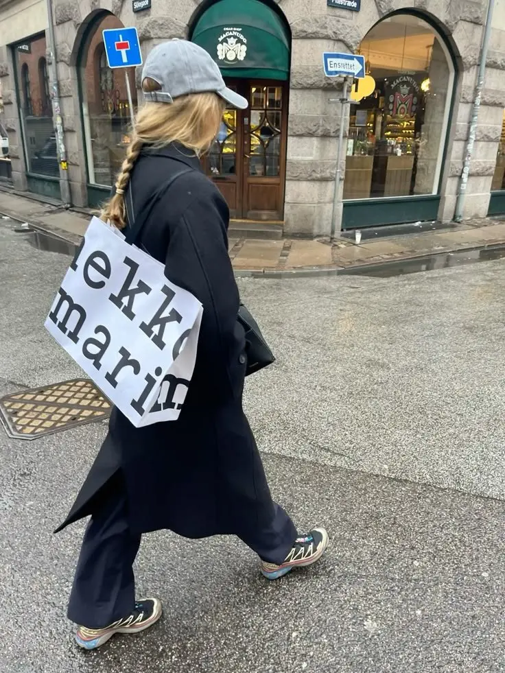 woman walking in long black wool coat flared black trousers grey baseball cap braid multicolor trail sneakers rainy street outfit 10