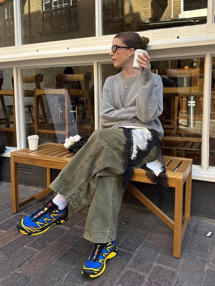 woman in grey knit sweater army green cargo pants white socks blue yellow salomon xt6 sneakers sitting on bench outside cafe outfit 12
