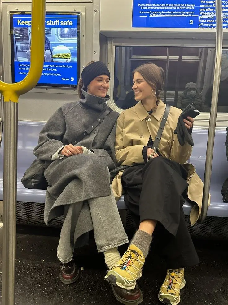 two women sitting on subway train wearing layered coats wide leg trousers one with mustard yellow salomon sneakers grey wool socks one with burgundy loafers outfit 13