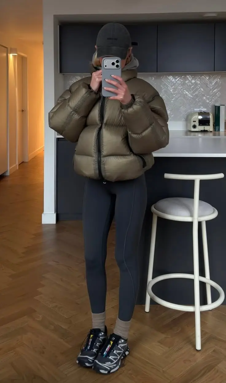 woman in oversized olive glossy puffer jacket charcoal leggings taupe socks dark grey baseball cap salomon trail runners kitchen mirror selfie outfit 14