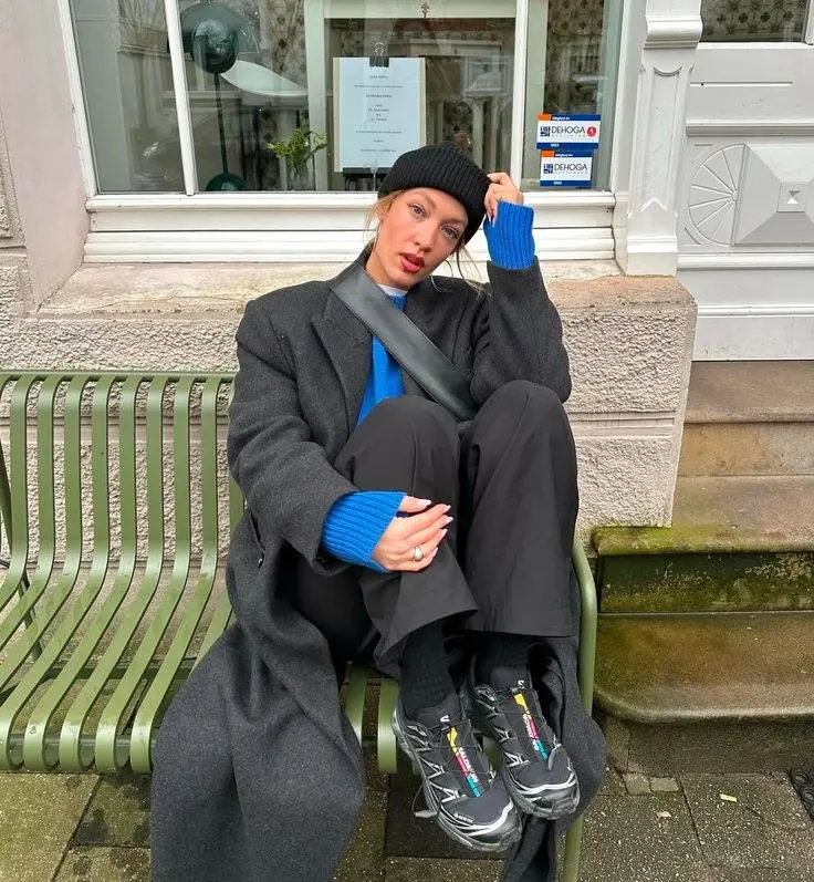 woman sitting on bench in long grey overcoat royal blue ribbed knit sweater black wide leg trousers black beanie black sling bag black salomon runners outfit 15