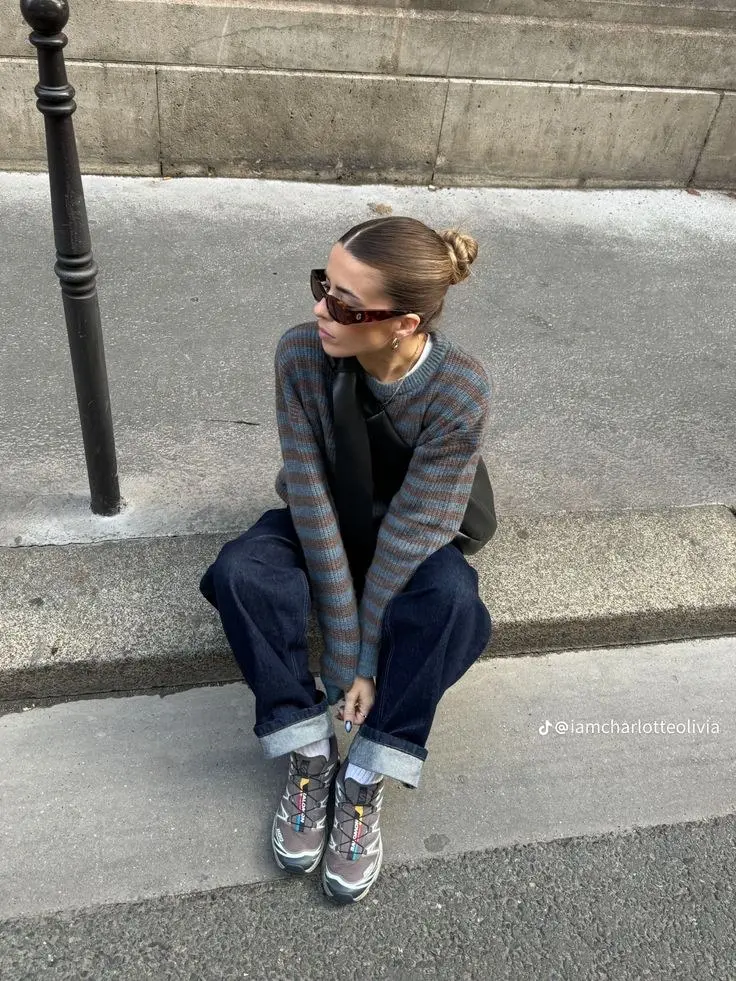 woman sitting on steps in striped brown grey knit sweater rolled dark indigo wide leg jeans black crossbody bag white socks grey brown salomon trail runners tortoiseshell sunglasses outfit 16