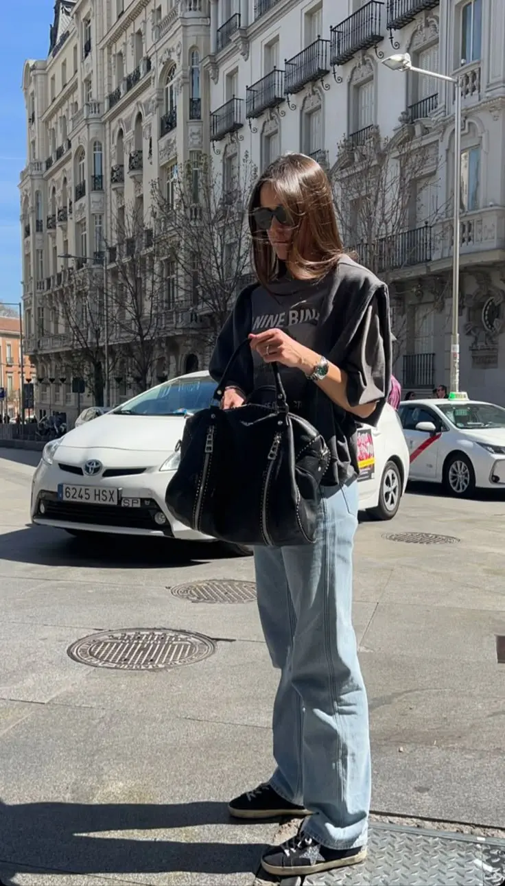 woman in black graphic sweatshirt light wash wide leg jeans black hardware bag and star sneakers street photo 1