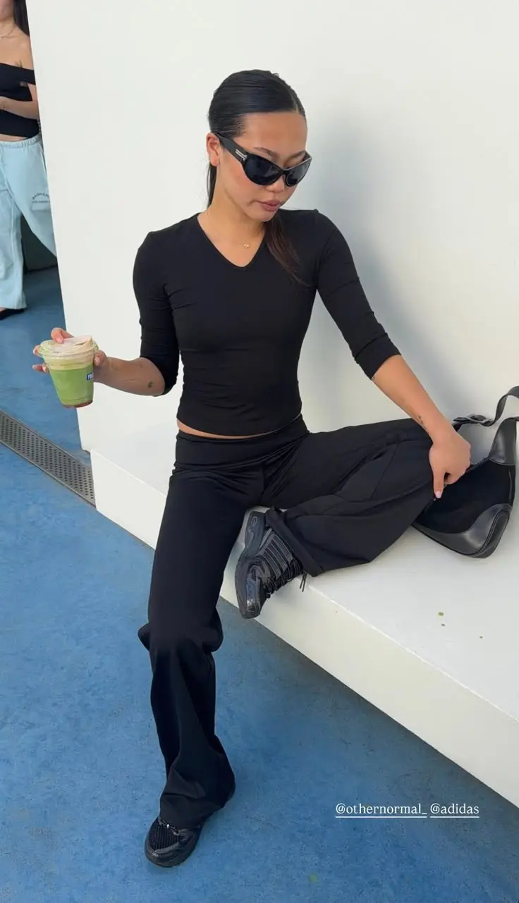 woman in all-black fitted three-quarter sleeve top wide leg black trousers black chunky adidas sneakers small black shoulder bag angular black sunglasses seated outdoor outfit 2