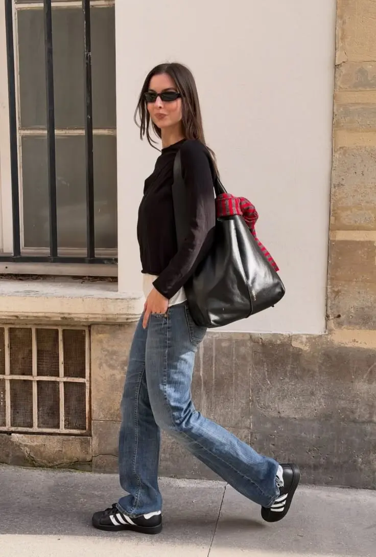 woman in black long sleeve fitted top white tee underneath medium wash straight leg jeans black adidas samba sneakers large black leather tote with red plaid scarf tied on handle small black sunglasses walking street outfit 4