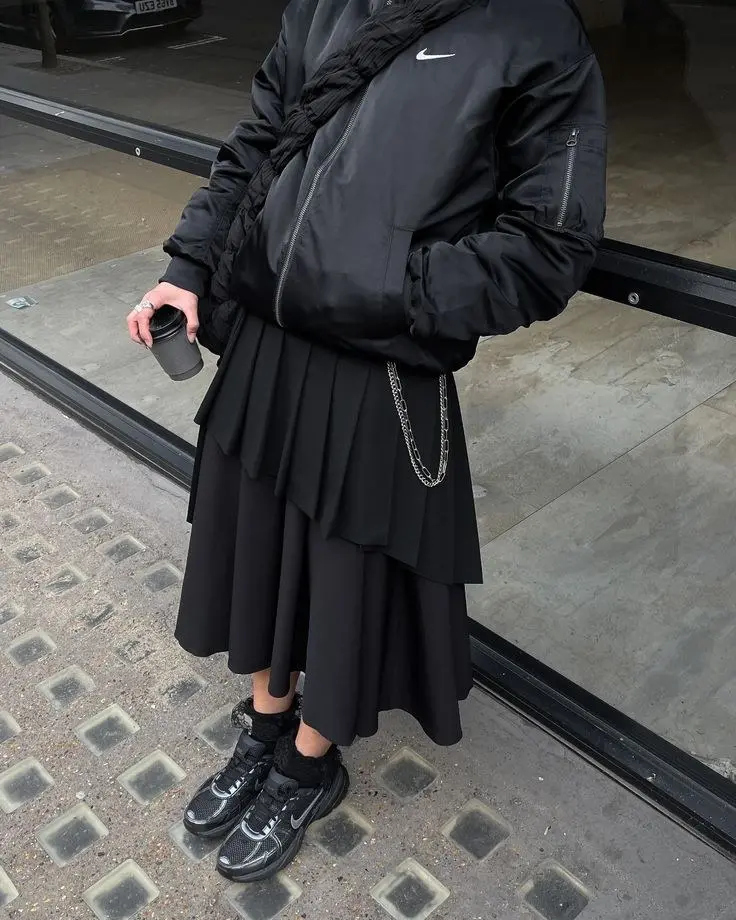 person in black oversized Nike puffer jacket black layered asymmetric pleated midi skirt chunky black Nike sneakers silver chain bag holding coffee street outfit 5