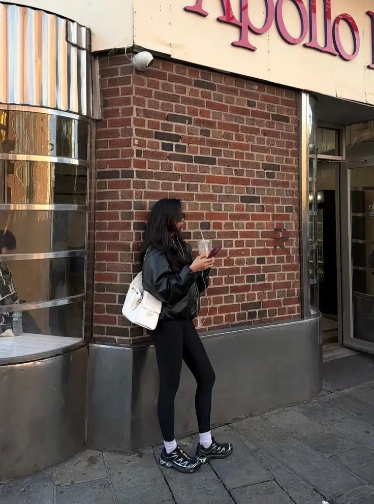 woman in black oversized faux leather jacket black leggings black trail sneakers white crew socks white structured backpack glasses leaning against brick wall outfit 6