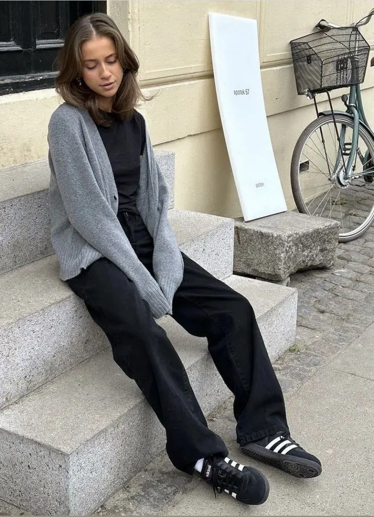 woman in grey oversized open cardigan black tee black wide leg jeans adidas samba black white sneakers white socks small gold earrings seated on stone steps outdoor outfit 7