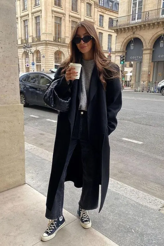 woman in long black wool overcoat grey crew neck sweater black wide leg jeans black converse chuck high top sneakers dark velvet shoulder bag small black sunglasses holding coffee cup street outfit 11