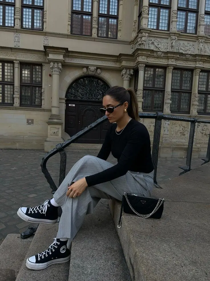 woman in fitted black long sleeve top grey plaid wide leg trousers black converse chuck high top platform sneakers white socks layered gold chain necklaces small black chain shoulder bag small sunglasses seated on stone outdoor outfit 12