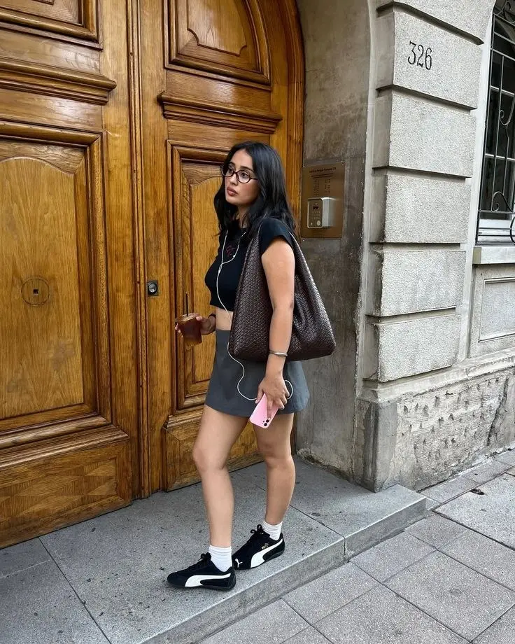 young woman in black sleeveless crop top grey athletic mini skirt black puma sneakers white crew socks large dark brown woven tote bag glasses earphones walking past ornate wooden door outdoor outfit 13