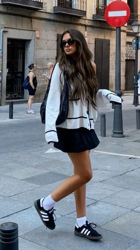 woman in white cardigan with black trim contrast detailing black pleated mini skirt adidas samba black gum sole sneakers white socks black quilted backpack large sunglasses wavy hair walking outdoor street outfit 14
