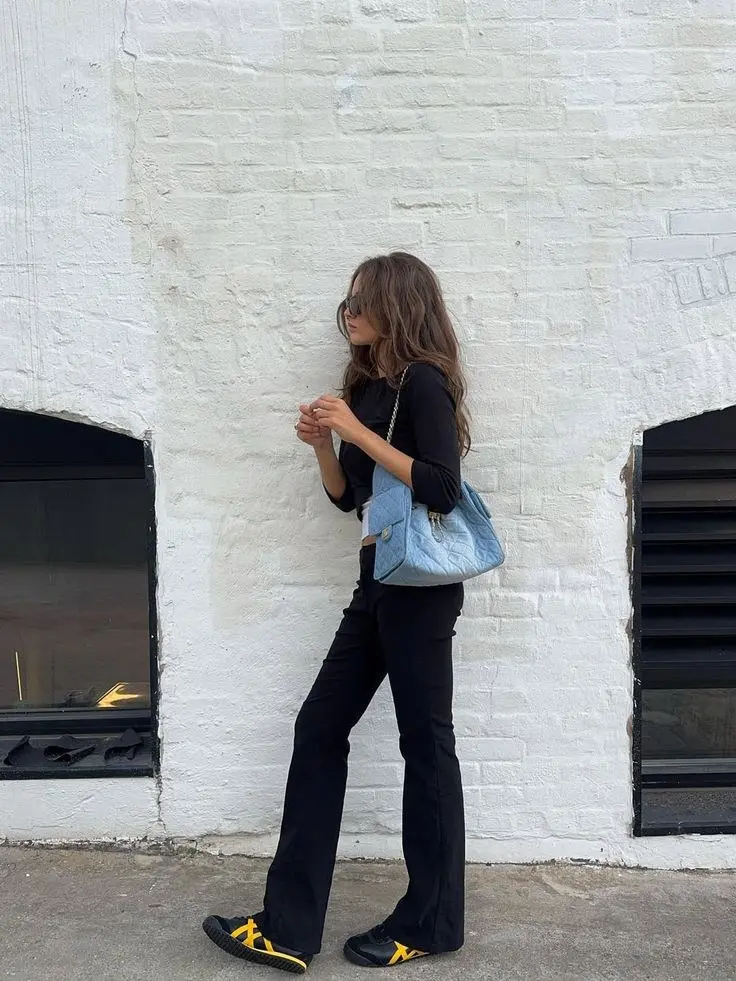 woman in black three quarter sleeve fitted top black flare leg trousers black yellow Onitsuka Tiger sneakers light blue denim quilted chain shoulder bag small sunglasses outdoor white brick wall outfit 16