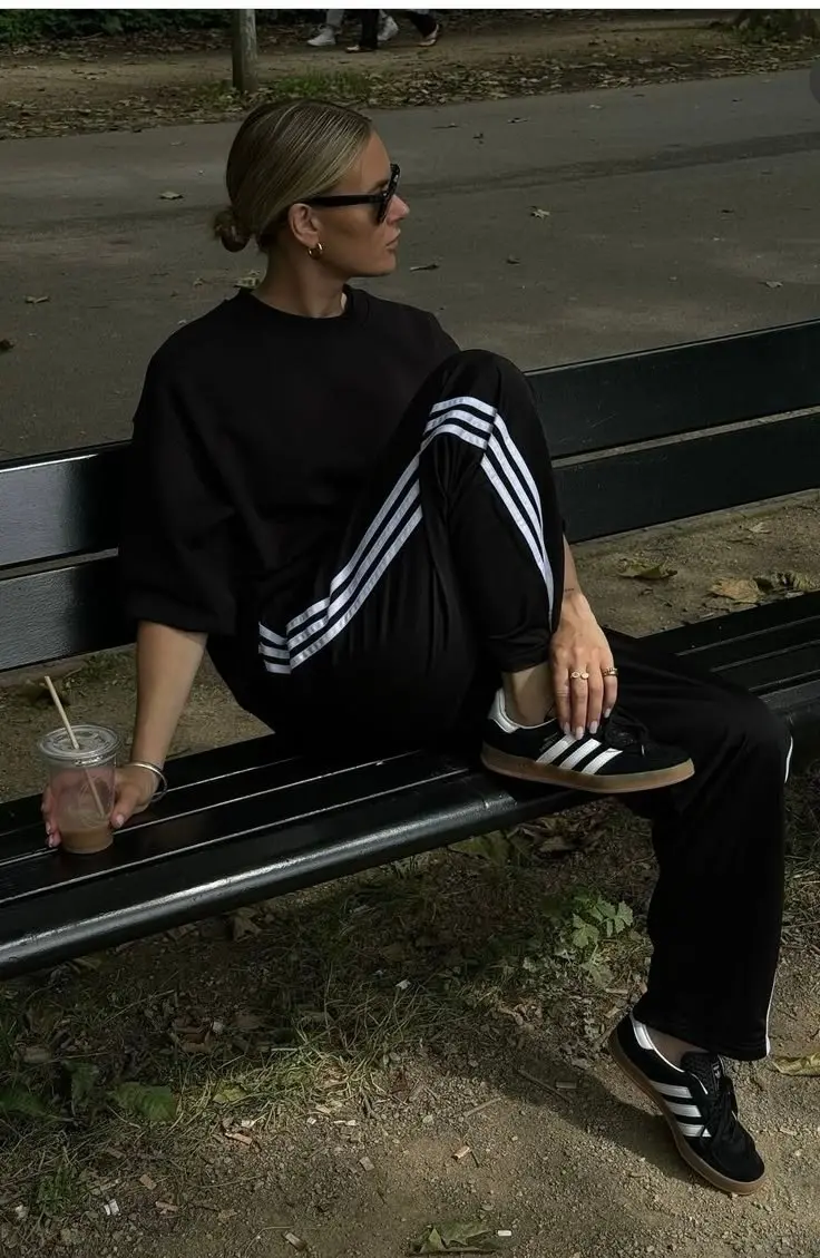 woman in all-black adidas oversized crewneck sweatshirt black three stripe flare track pants adidas samba gum sole black sneakers gold hoop earrings slicked bun sunglasses park bench iced coffee outfit 20