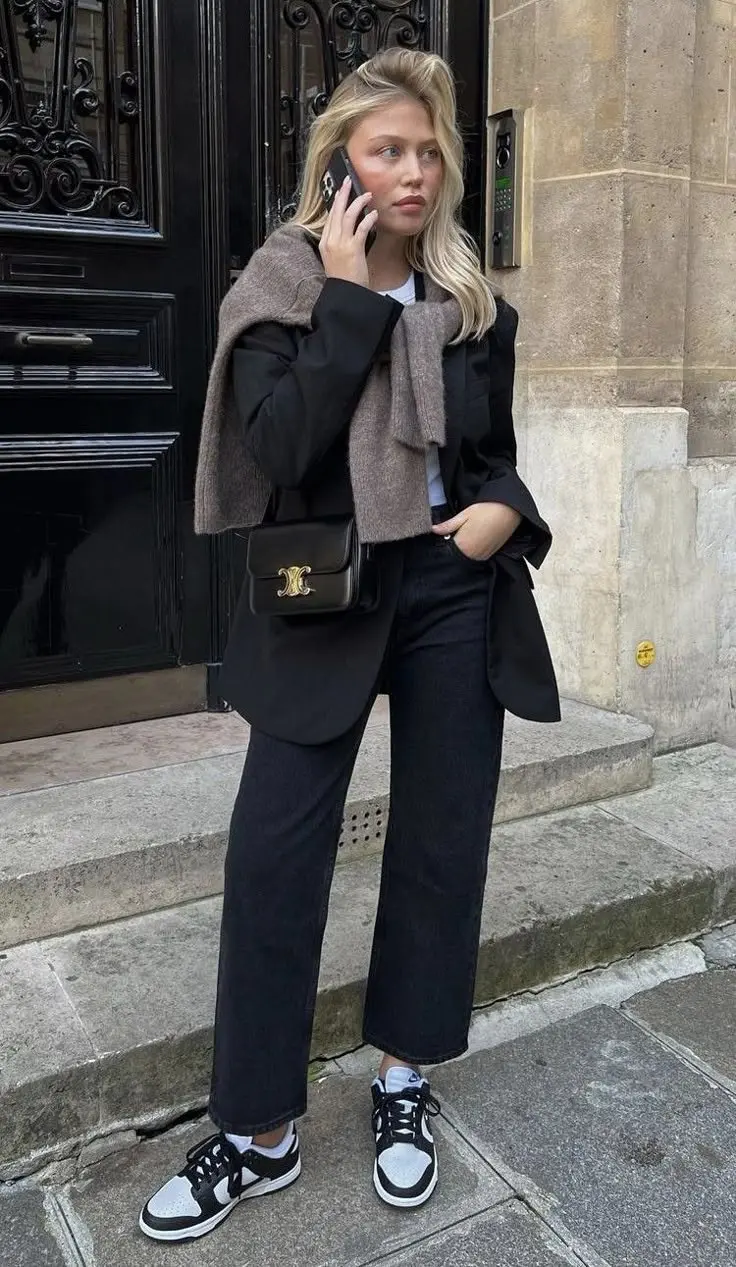 woman in black oversized blazer beige knit sweater tied at shoulders white tee straight black jeans black white Nike Dunk sneakers white socks small black chain crossbody bag wavy blonde hair phone call outdoor street outfit 21