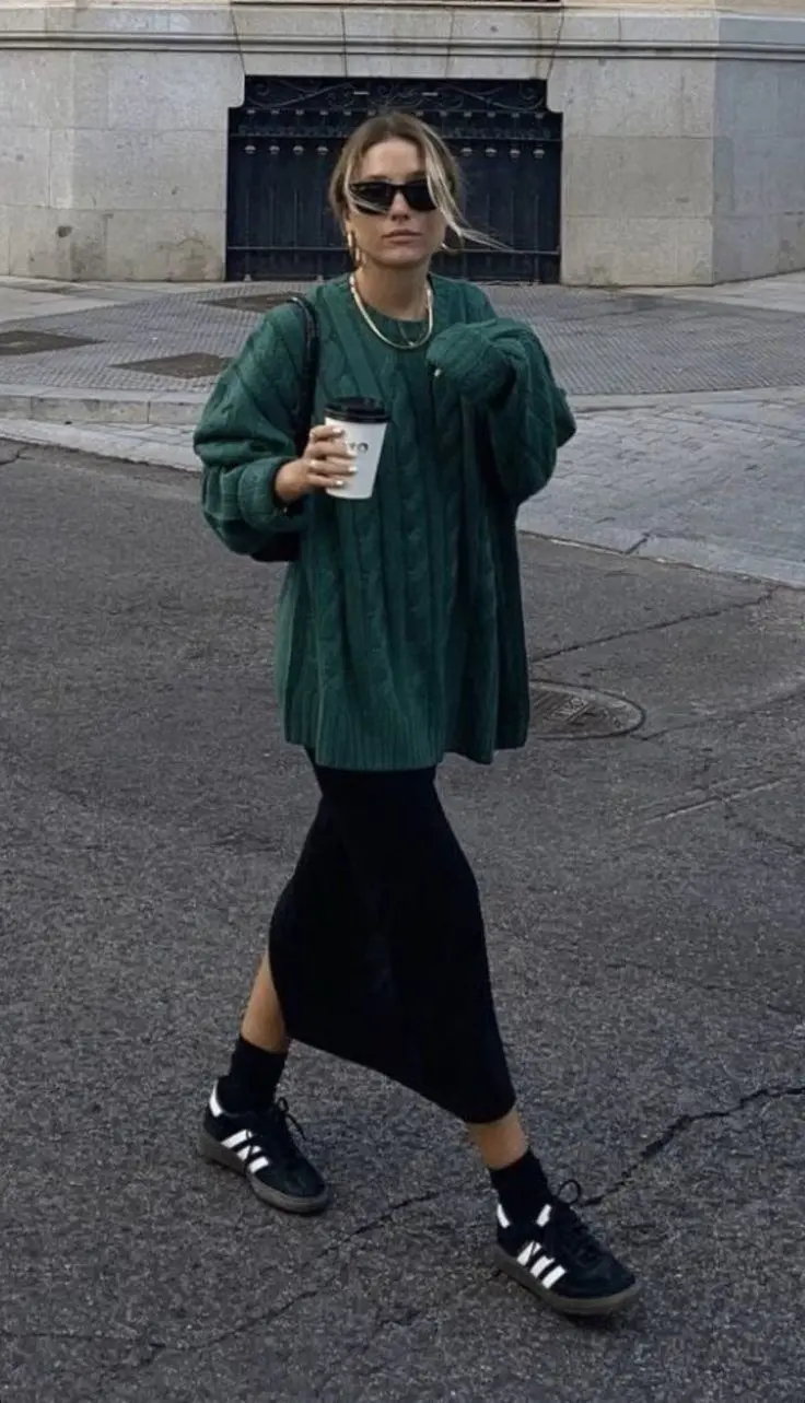 woman in oversized forest green cable knit sweater black satin midi slip skirt black crew socks adidas samba black gum sole sneakers gold layered chain necklaces small sunglasses walking outdoor street outfit 22