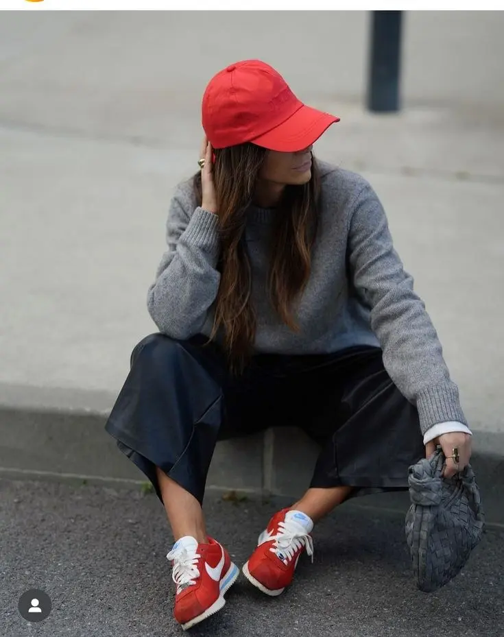 woman wearing red baseball cap grey knit sweater dark leather trousers red Nike Cortez sneakers woven grey bag street style outfit 1