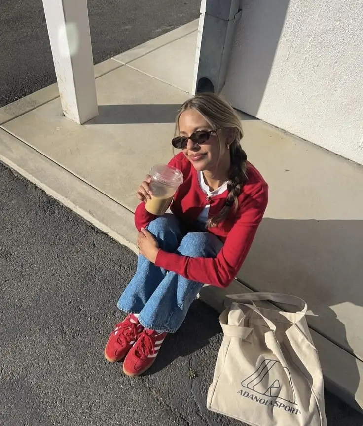 woman in red cardigan white tee light wash jeans red Adidas Samba sneakers cream canvas tote sunglasses braided pigtails sitting outdoors outfit 2