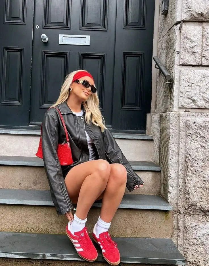 blonde woman in oversized black leather jacket white tee mini shorts red Adidas Gazelle sneakers white socks red mini bag red headband sunglasses sitting on steps outfit 6