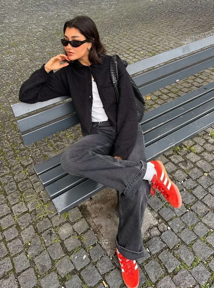 woman in black textured cardigan white tee grey wide leg jeans red Adidas Samba sneakers white socks woven black bag sunglasses sitting on bench outdoors outfit 7