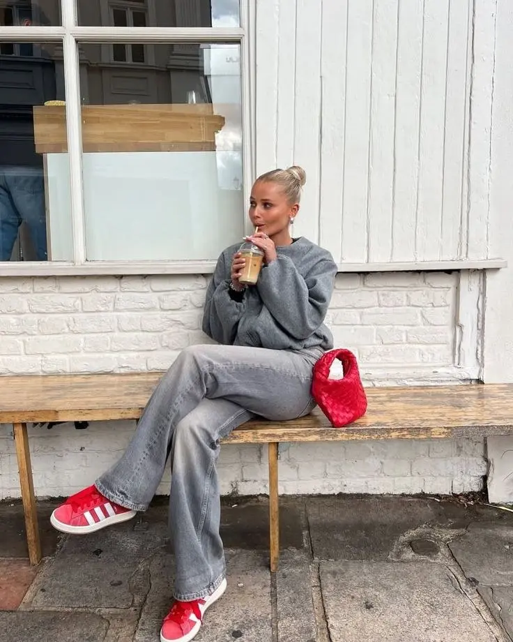 blonde woman in grey oversized sweatshirt grey wide leg flared jeans red Adidas Samba sneakers red woven pouch bag hair in bun sitting on bench outside cafe outfit 9
