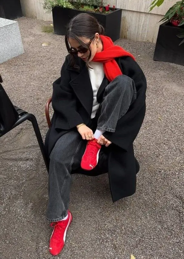 woman in black oversized long coat white sweatshirt grey wide leg jeans red knit scarf red Puma sneakers sitting on chair outdoors sunglasses gold jewelry outfit 12