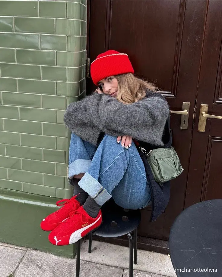 young woman in red beanie grey oversized chunky knit cuffed light blue jeans red Puma sneakers dark socks olive green crossbody bag sitting against tiled wall outfit 13