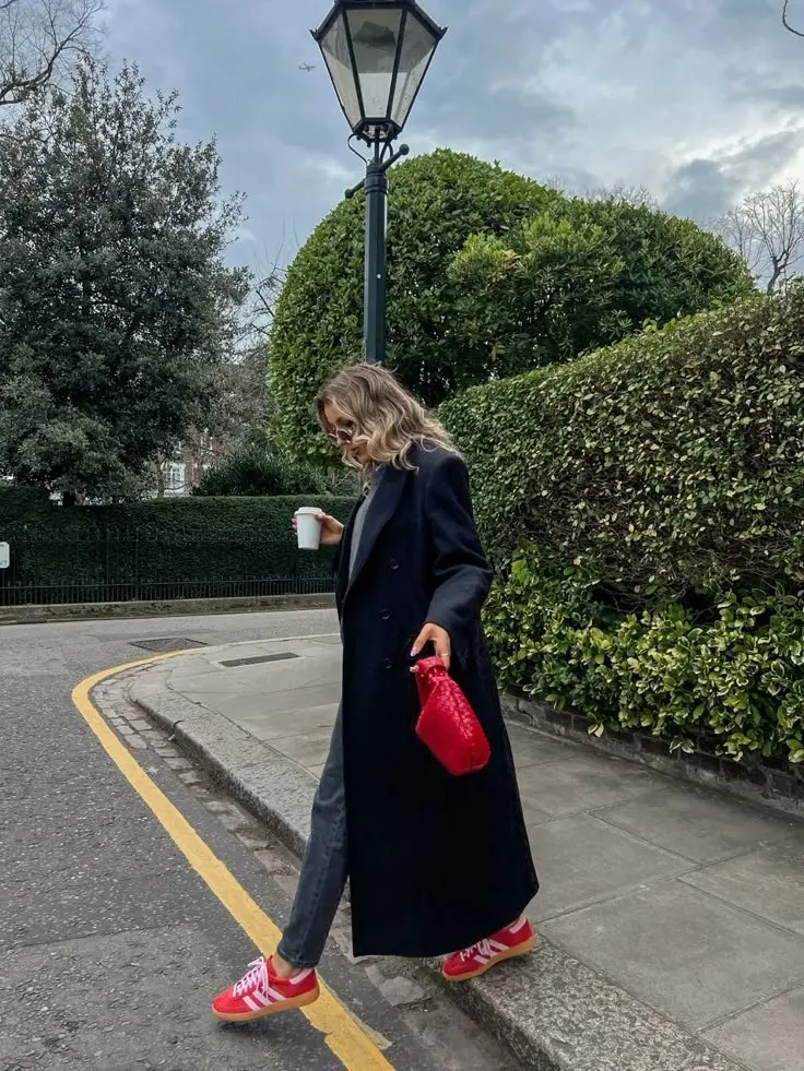 woman in navy double breasted maxi coat grey slim jeans red Adidas Samba sneakers red woven pouch bag wavy hair walking on street holding coffee cup outfit 15