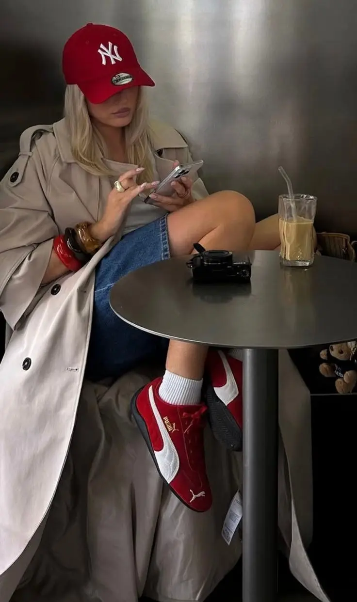 blonde woman in red NY baseball cap beige trench coat white tee denim mini skirt red Puma sneakers white socks sitting at cafe table with bangles and rings outfit 16