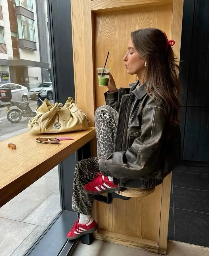 woman in distressed dark leather jacket leopard print wide leg trousers red Adidas Samba sneakers white socks red hair claw clip sitting by cafe window with cream bag and green matcha outfit 18