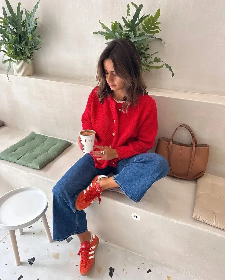 woman in red chunky knit cardigan white tee wide leg blue crop jeans red Adidas Samba sneakers tan leather tote bag pearl earrings gold necklaces sitting indoors with coffee outfit 19