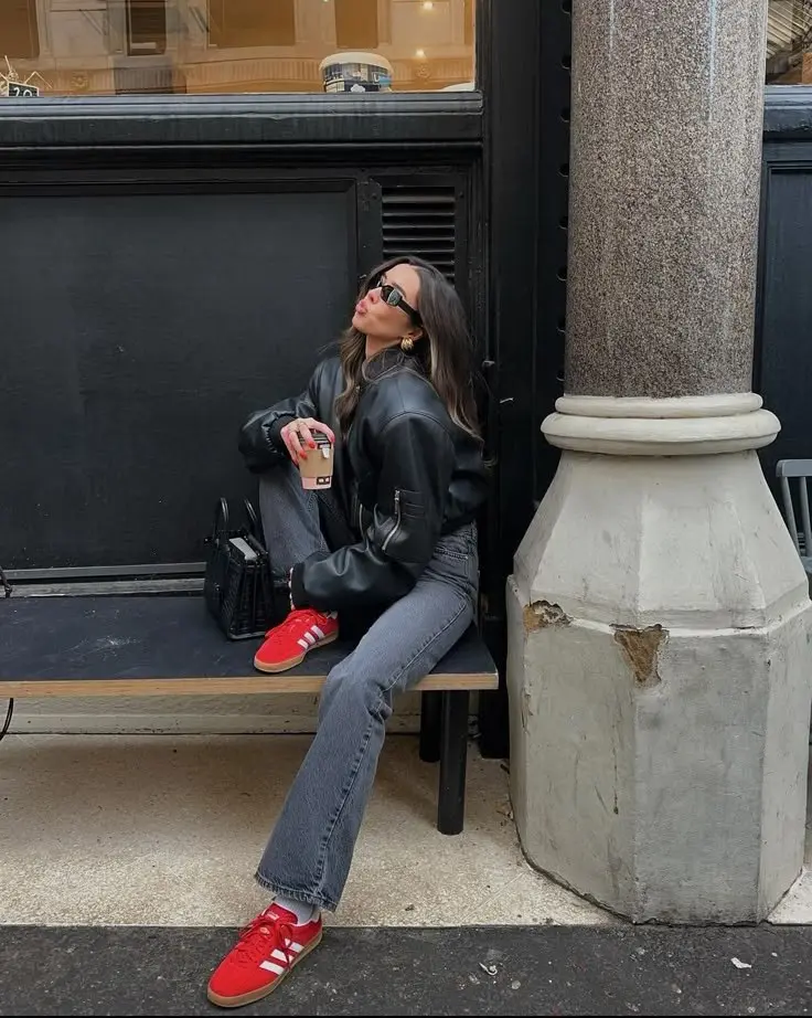 woman in black oversized leather bomber jacket grey straight leg jeans red Adidas Samba sneakers white socks small black structured bag gold hoop earrings sunglasses sitting on bench street style outfit 22
