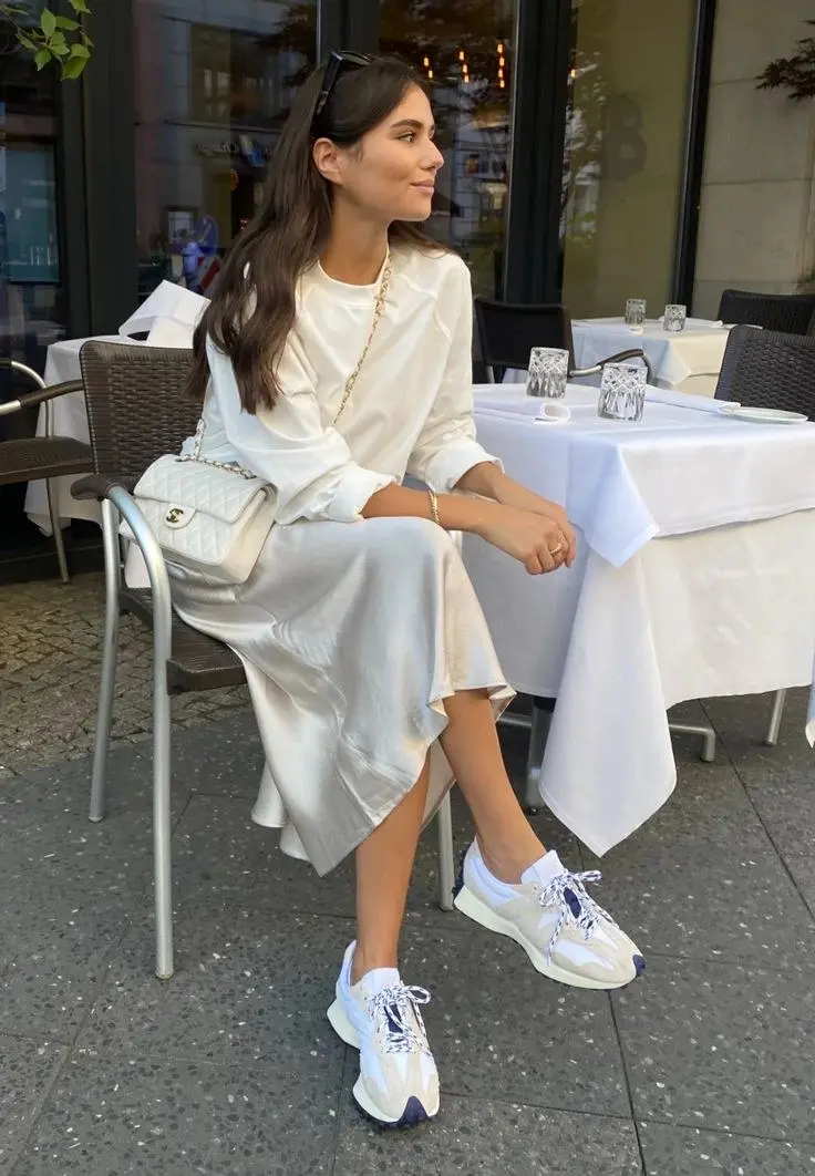 woman in white top champagne satin midi skirt white New Balance sneakers white quilted chain bag seated at outdoor restaurant outfit 4