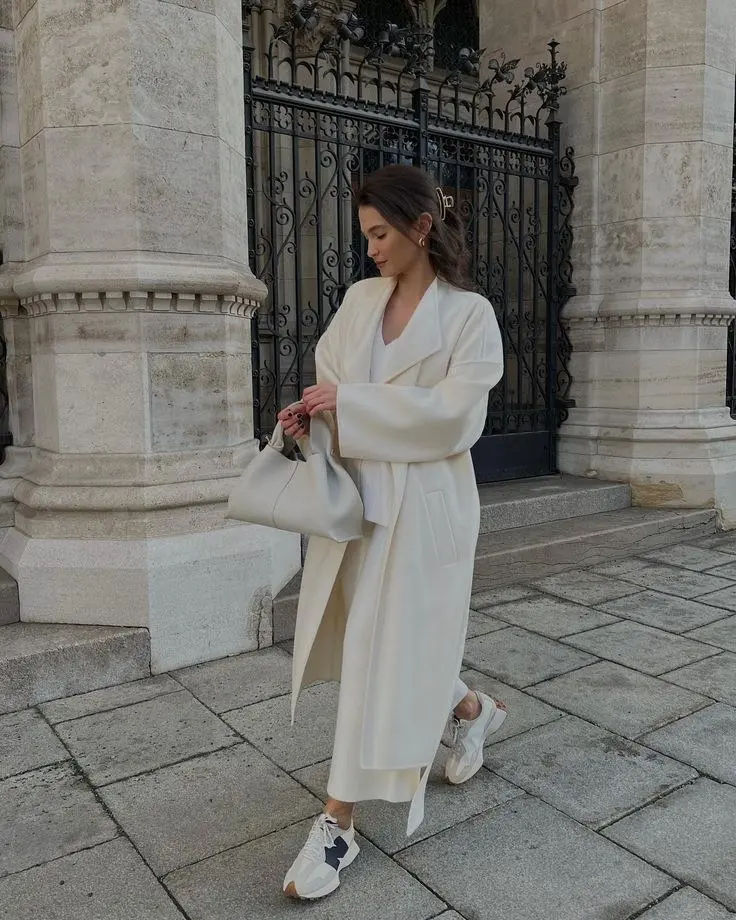 woman in long ivory robe coat white midi skirt soft grey shoulder bag cream New Balance sneakers gold earrings hair clip outdoor architecture outfit 8