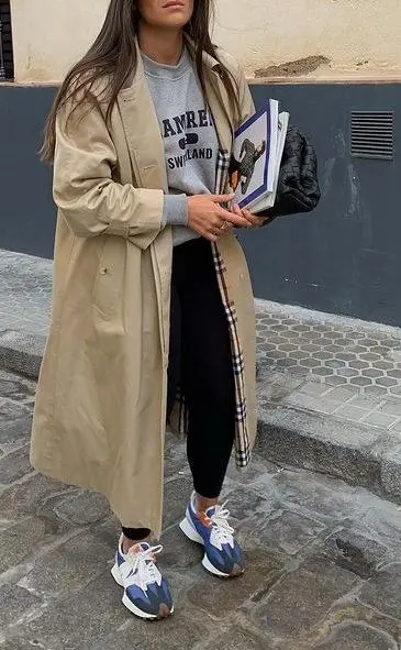 woman in camel trench coat grey graphic sweatshirt black leggings blue orange New Balance sneakers holding books and black bag outdoor outfit 12