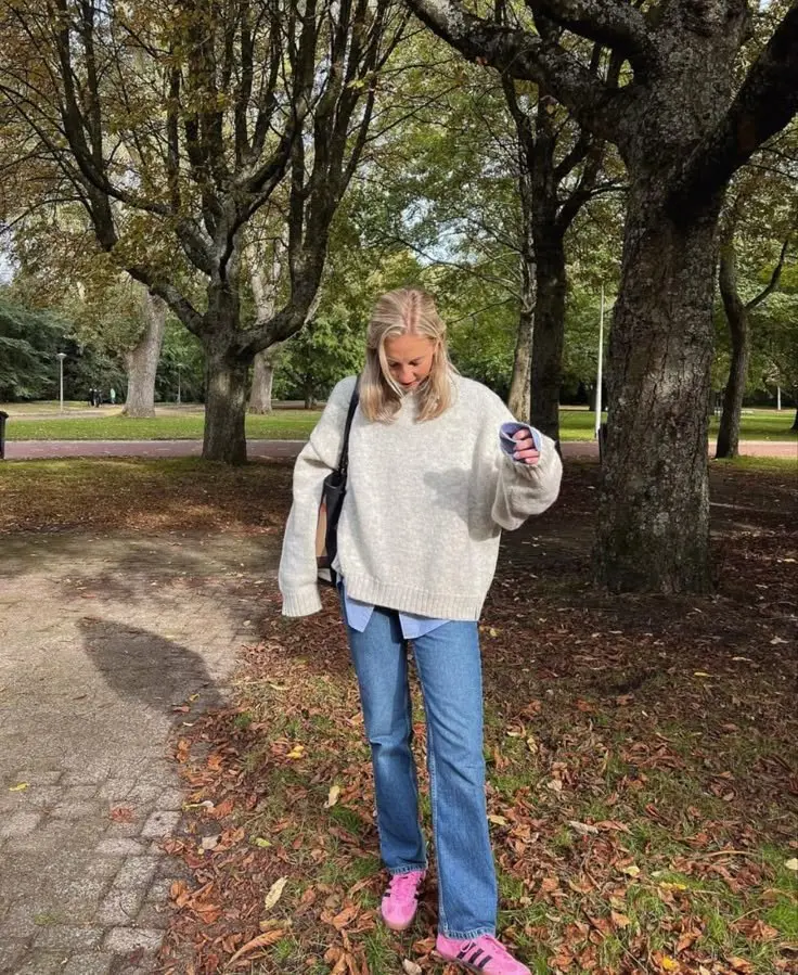 woman outdoors in cream oversized knit sweater layered over blue oxford shirt, straight medium-wash jeans, bright pink sneakers, small black crossbody bag, photo 5