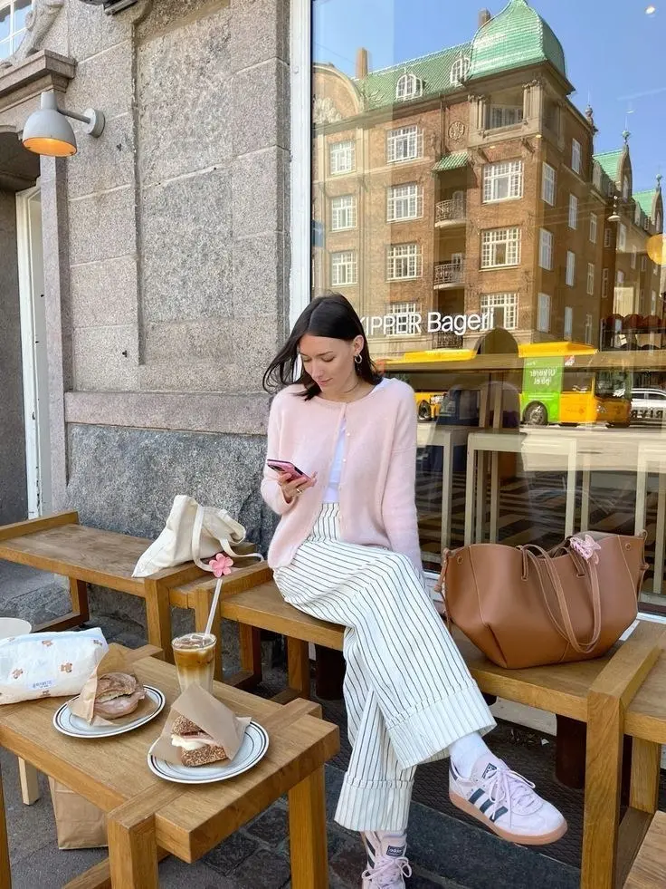 woman seated outside cafe in pink fuzzy cardigan over white tank, white and cream vertical-stripe wide-leg trousers, pink sneakers with white socks, large cognac tote bag, pink flower straw, photo 13