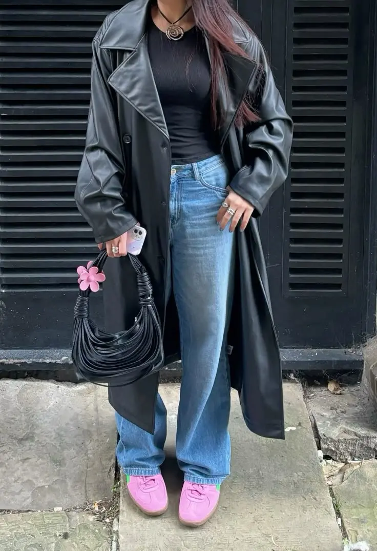 woman in long black double-breasted leather trench coat, black ribbed fitted top, straight mid-wash jeans, bright pink sneakers, black knotted bag with pink flower charm, silver rings, photo 15