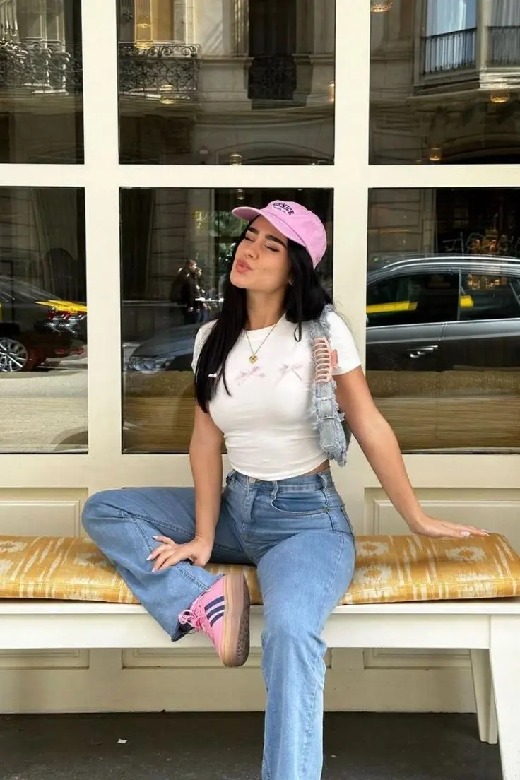 woman seated on bench in pink velvet baseball cap, white bow graphic crop tee, light-wash baggy jeans, pink sneakers, distressed denim vest over shoulders, gold necklace, photo 17