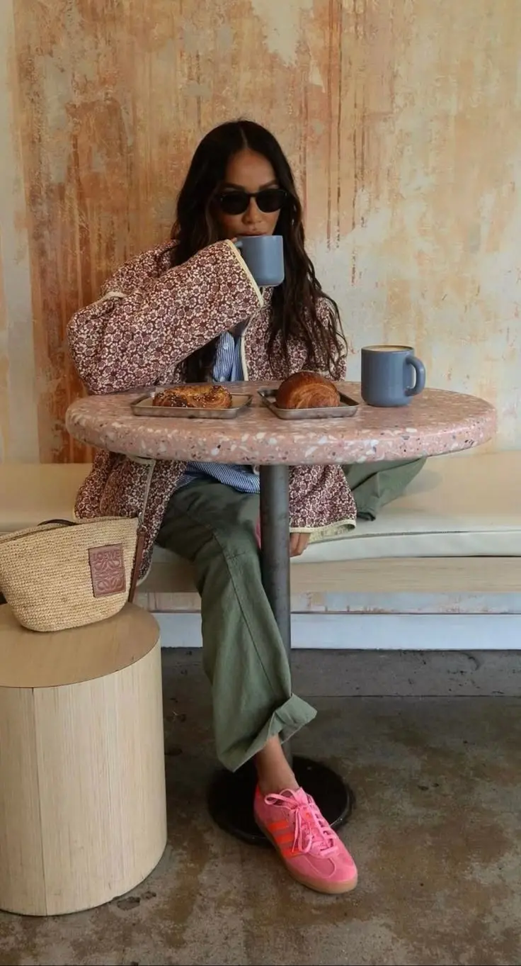 woman seated at terrazzo cafe table in floral cream and dark red quilted jacket, blue striped shirt underneath, olive cargo trousers, coral pink sneakers, raffia bag with leather label, sunglasses, photo 18