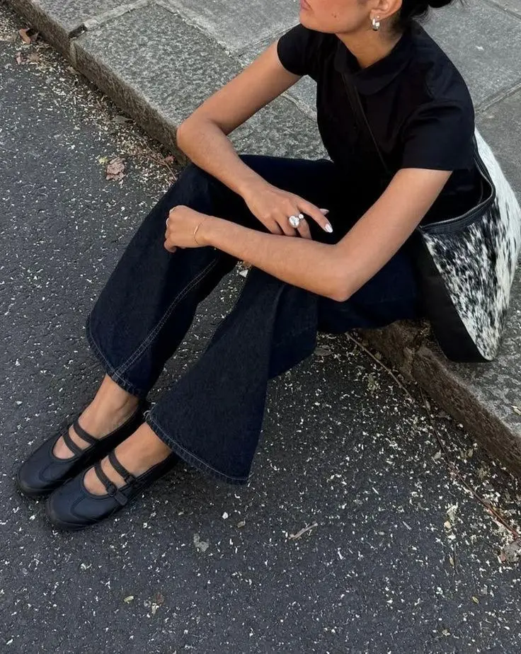 woman seated on curb in black tee dark indigo wide-leg jeans black double-strap Mary Jane sneakers cowhide pattern tote bag silver earrings outfit photo 2