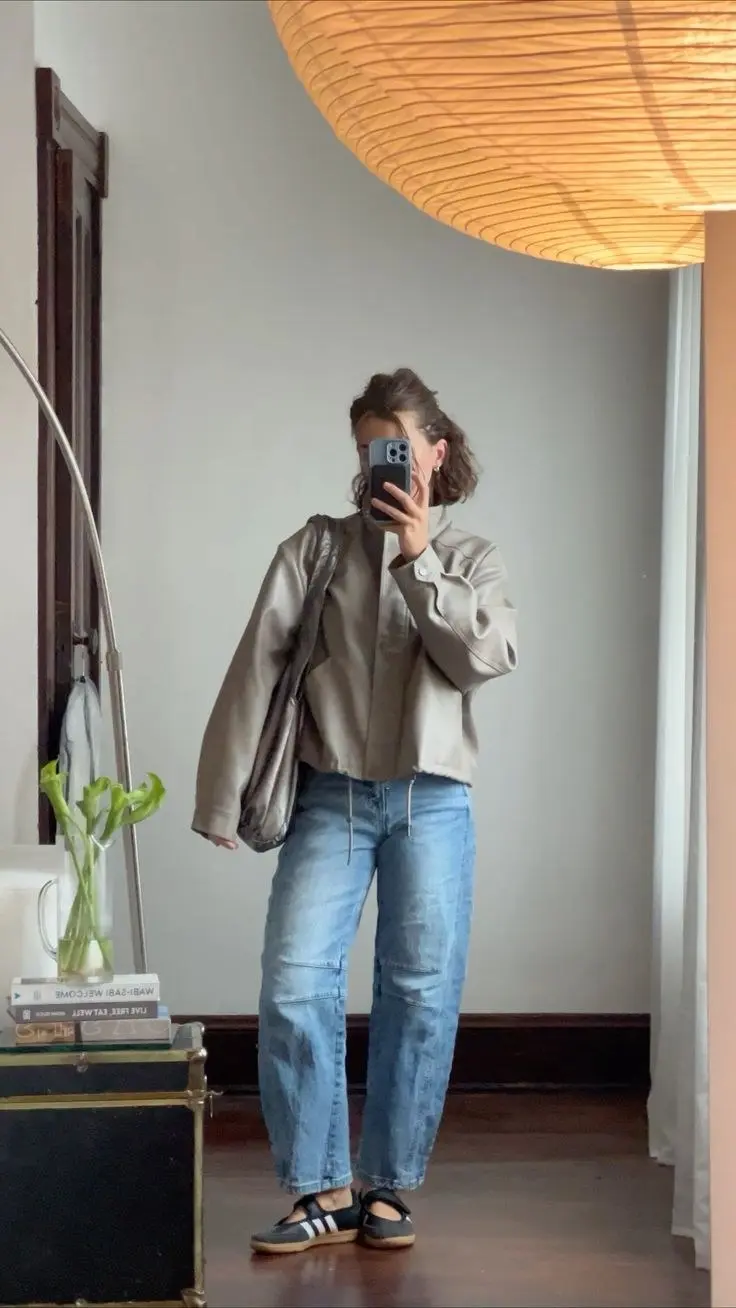 woman taking mirror selfie in taupe cropped faux leather jacket light wash barrel drawstring jeans black sport strap flat shoes indoor outfit photo 4