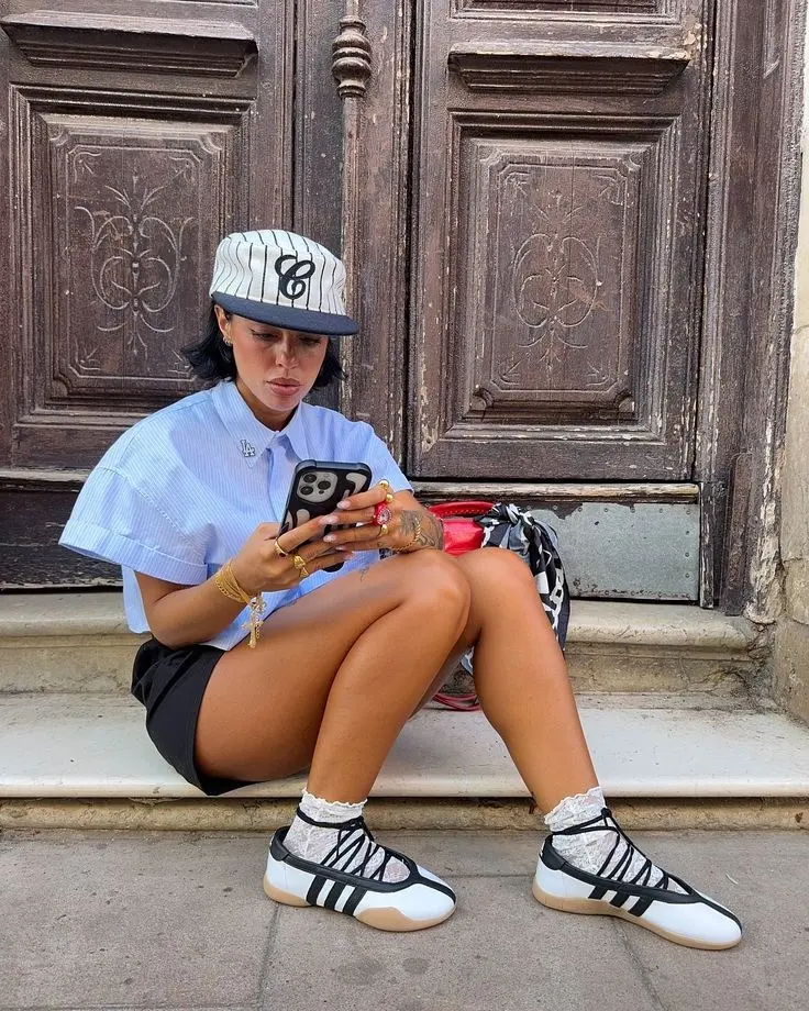 woman seated on steps in blue Oxford crop shirt black bike shorts pinstripe baseball cap lace ankle socks black white ballet sneakers gold jewelry photo 5