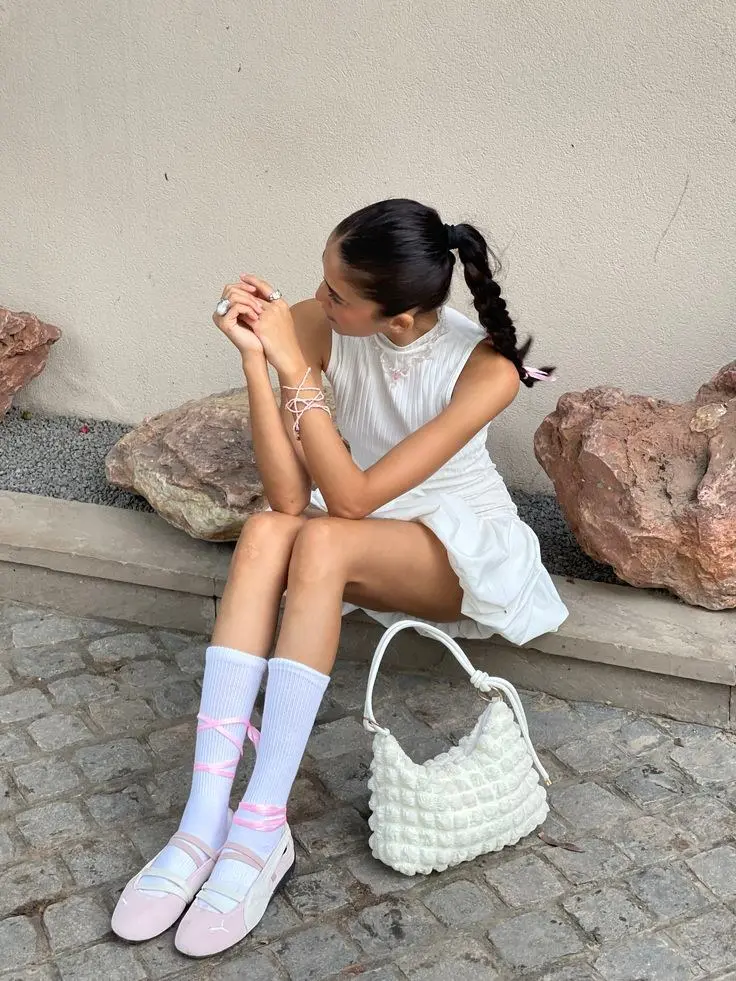 woman seated on stone bench in white ribbed sleeveless mini dress white crew socks with pink ribbon ties pink ballet sneakers white quilted bubble bag braided ponytail with ribbon photo 6