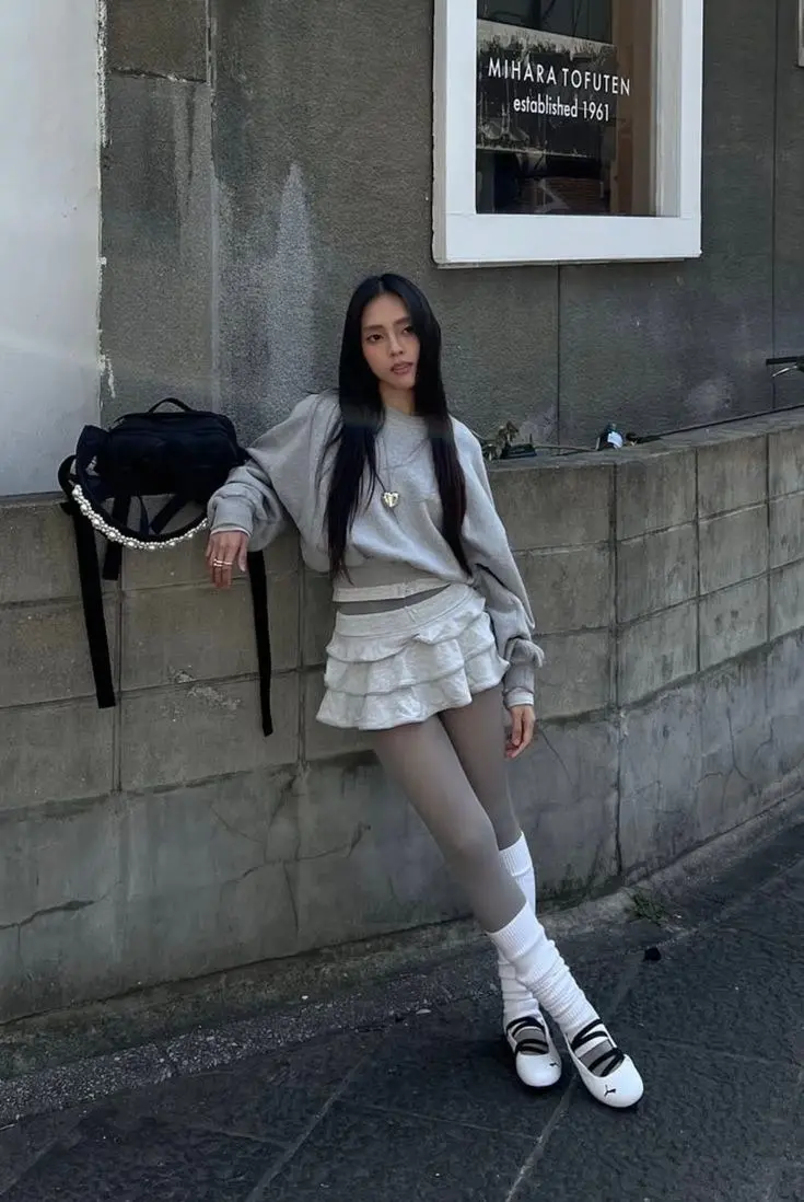 woman leaning against wall in grey oversized sweatshirt white tiered ruffle mini skirt sheer grey tights white slouch socks white Mary Jane ballet sneakers gold heart necklace black pearl chain bag photo 7