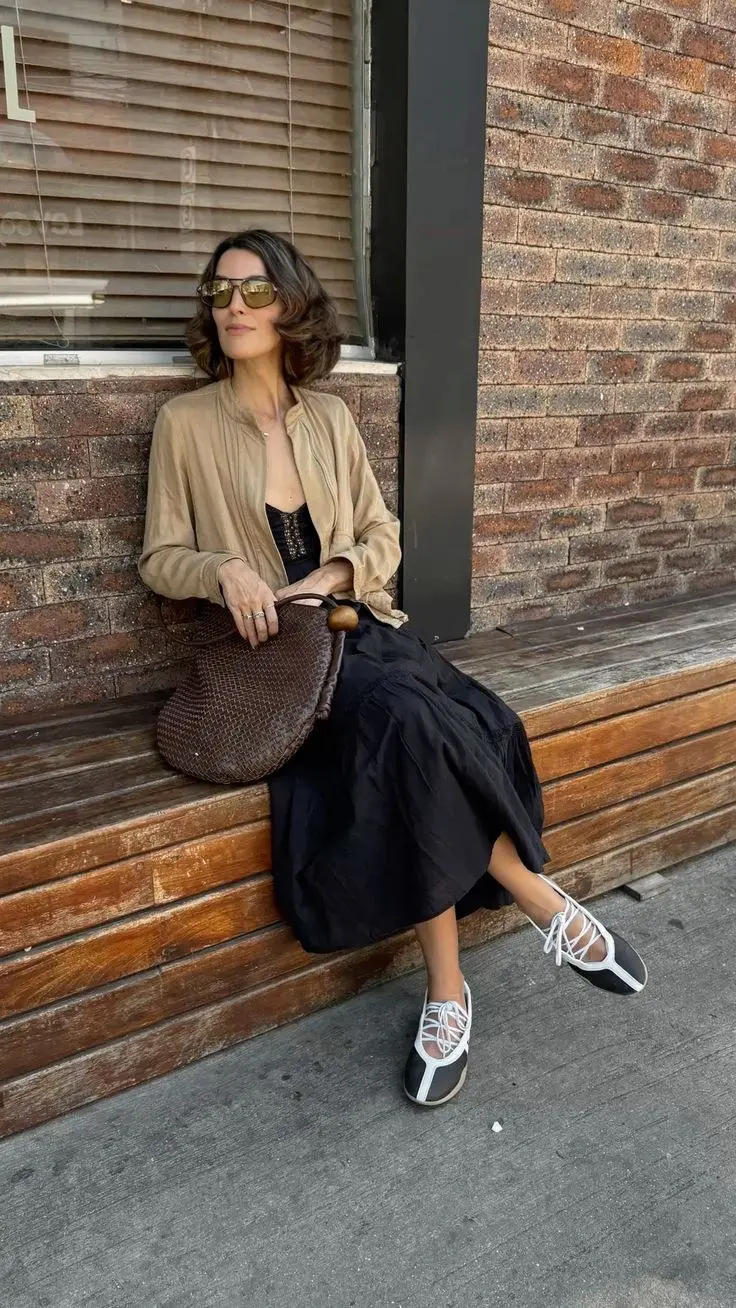 woman seated on wooden bench in camel suede bomber jacket black lace cami black fluid midi skirt white lace-up sport ballet sneakers dark woven leather half-moon bag tinted sunglasses photo 8