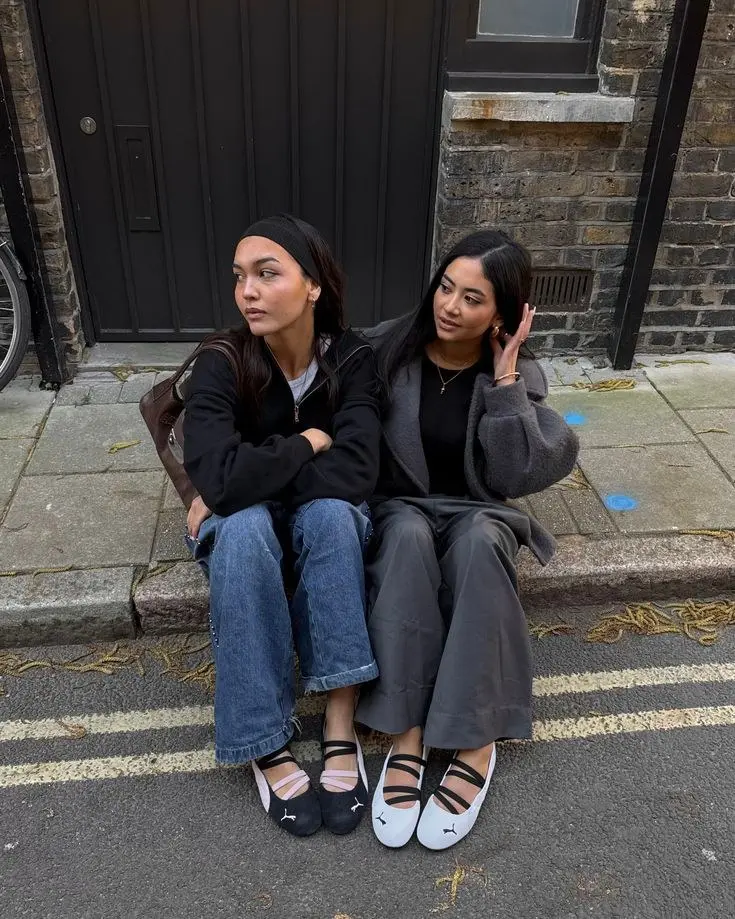 two women seated on street curb one in black zip fleece wide-leg jeans brown bag one in grey wool coat black turtleneck grey trousers both wearing Puma ballet sneakers photo 9