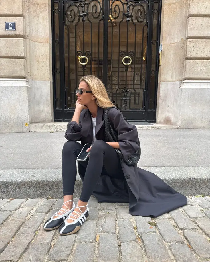 woman seated on cobblestone street in long charcoal tailored coat white tee black leggings white lace-up ballet sneakers dark woven clutch bag cat-eye sunglasses photo 10