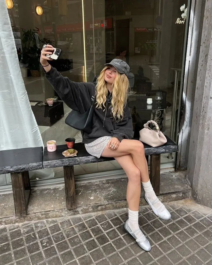 woman seated outdoors in oversized dark grey hoodie silver sequin mini skirt white crew socks silver white ballet sneakers bucket bag crossbody blush tote bag baseball cap photo 11