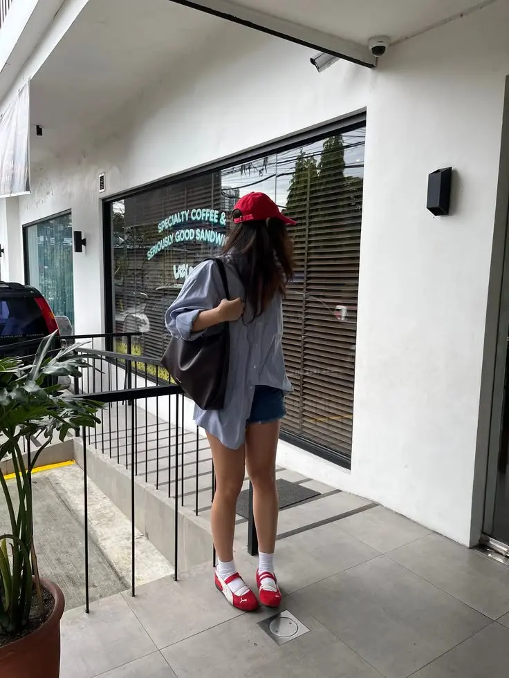 woman shot from behind in light blue oversized shirt denim shorts red baseball cap white crew socks red Mary Jane ballet sneakers large dark brown tote bag street photo 12
